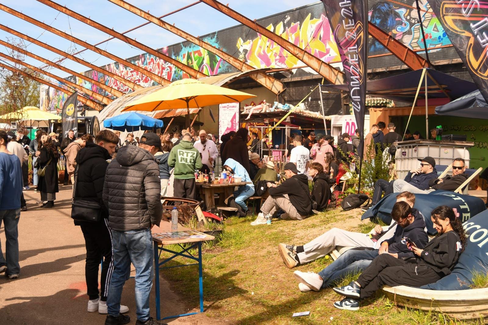 Druk buitenterrein van Hal 25 met parasols, een graffitimuur en bezoekers op het gras